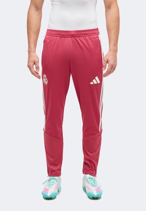 Mand iført røde Adidas træningsbukser med hvide striber og logoer, kombineret med hvide og turkise fodboldstøvler, stående mod en hvid baggrund.