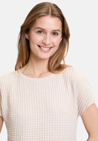 Light beige textured top featuring a grid pattern, round neckline, and short sleeves. Soft fabric with a subtle sheen.