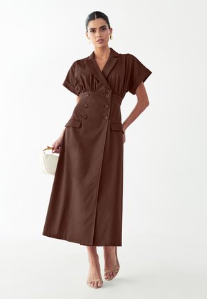 Vrouw draagt een bruin double-breasted midijurk met korte mouwen, houdt een witte handtas vast en draagt beige open-toe hakken, tegen een witte achtergrond.