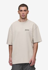 Beige ylisuuri t-paita, joka on valmistettu puuvillasta. Paidassa on pyöreä kaula-aukko ja lyhyet hihat, sekä pieni musta grafiikka edessä.
