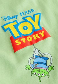 Disney Pixar Toy Story-Logo über einem grünen dreiäugigen Alien-Spielzeug, das in einer Greifarm-Kralle auf hellgrünem Stoff gefangen ist.