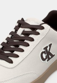 Αθλητικά παπούτσια Calvin Klein με επάνω μέρος από κρέμα σουέντ, καφέ κορδόνια και λαστιχένια σόλα. Διαθέτουν έντονο λευκό λογότυπο.