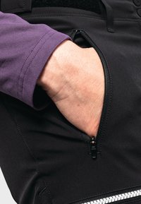 Schwarze Reißverschlusstasche an der Hose, Hand steckt in der Tasche. Das Material ist glatt mit einer matten Oberfläche. Lila Ärmel im Hintergrund sichtbar.