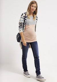 Conjunto de maternidade com um top em camadas em tons de cinza e pêssego, jeans de ganga slim, um cardigã com padrão azteca e ténis cinzas.