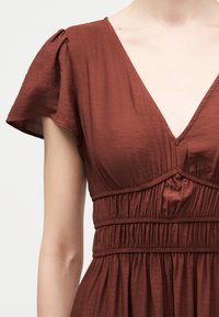 Γυναίκα που φοράει υφασμάτινο φόρεμα σε χρώμα σκουριάς καφέ με κοντά φουσκωτά μανίκια, λαιμόκοψη V και ζώνη με ραντάκια στη μέση.
