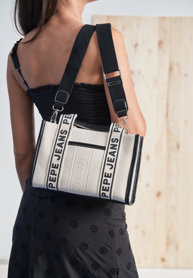 Femme portant un haut noir et une jupe tient un sac crème Pepe Jeans avec des poignées noires et des lanières avec logo sur son épaule.