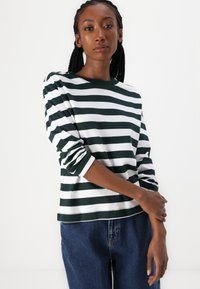 Camicia a maniche lunghe a righe verdi e bianche in un tessuto morbido, con scollatura rotonda e polsini arrotolati, abbinata a pantaloni in denim blu.