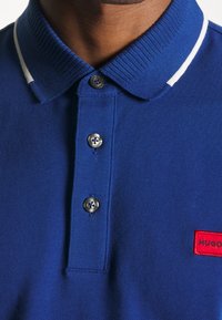 Gros plan d'une personne portant un polo bleu avec des rayures blanches sur le col et trois boutons, avec une étiquette rectangulaire rouge sur la poitrine.