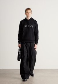 Μαύρο φούτερ με κουκούλα και λογότυπο «BOSS», συνδυασμένο με φαρδιά μαύρη παντελόνα. Τα αξεσουάρ περιλαμβάνουν μία μαύρη τσάντα και παπούτσια, τοποθετημένα σε ουδέτερο φόντο.