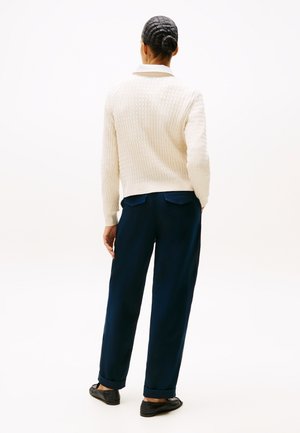 Kobieta z plecionym kokiem ubrana w biały sweter o fakturze, granatowe podwinięte spodnie i czarne mokasyny, stojąca tyłem na jasnym tle.