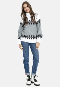 Kobieta ubrana w szary i biały sweter z golfem z wzorem, niebieskie podwinięte jeansy oraz białe, sznurowane buty z czarną podeszwą i detalami.