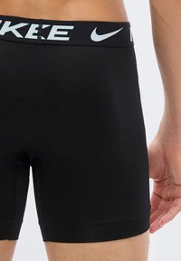 Κοντινό πλάνο ενός ατόμου που φοράει μαύρα σορτς Nike με ορατό λογότυπο στη μέση και μέρος του χεριού του να ακουμπά δίπλα στον μηρό.