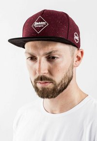 Bordó sapka texturált anyagból, fehér gyémánt logóval, fekete peremmel, sima fekete alsó résszel, egy sima fehér pólót viselő személy által viselve.