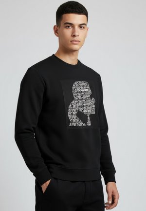 Młody mężczyzna ubrany w czarną bluzę z kapturem z białą grafiką w kształcie sylwetki, stworzoną z powtarzającego się tekstu „Karl Lagerfeld”, stojący na jasnym tle.