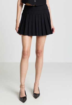 Donna che indossa una minigonna nera plissettata, tacchi neri a punta e una cavigliera dorata su sfondo semplice.