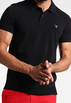 Mężczyzna w czarnej koszulce polo GANT i czerwonych spodniach, z założonymi z przodu rękami, na jasnym, jednolitym tle.