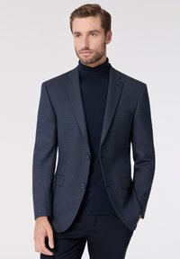 Navyblauer Blazer mit Textur, zwei Knöpfen, Schlapelkragen und Fronttaschen, kombiniert mit einem navyblauen Rollkragensweater. Taillierte Passform.