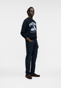 Μπλε ναυτικό φούτερ με λευκό γραφικό βουνού και κείμενο, συνδυασμένο με σκούρο τζιν και καφέ παπούτσια, σε απλό ανοιχτό φόντο.