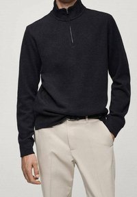 Uomo che indossa un maglione nero con zip a maniche lunghe infilato in pantaloni beige chiaro con una cintura nera, con una mano in tasca.