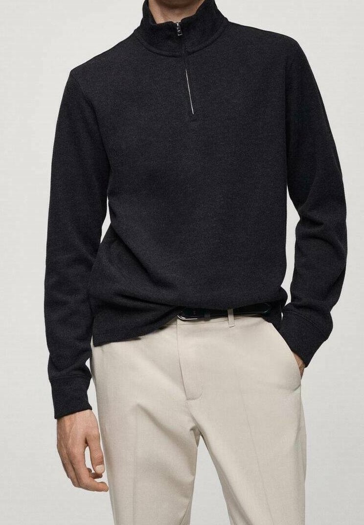 Uomo che indossa un maglione nero con zip a maniche lunghe infilato in pantaloni beige chiaro con una cintura nera, con una mano in tasca.