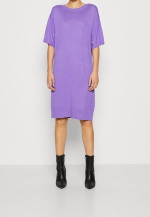 Kobieta w luźnej, sięgającej kolan, fioletowej sukience z krótkimi rękawami, w czarnych botkach do kostek, stojąca na jednolitym jasnym tle.