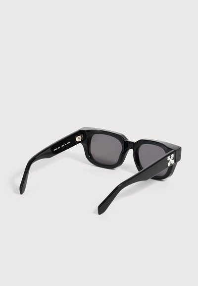 Lunettes de soleil rectangulaires noires avec des montures épaisses et des verres foncés, présentant un logo blanc sur la branche, sur un fond clair uni.