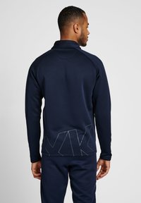 Pull-over de sport bleu marine avec un col haut, des manches longues et un motif textural géométrique au bas du dos. Tissu doux.