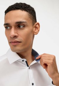 Camicia bianca con colletto strutturato, rivestimento interno blu a contrasto e bottoni scuri. Il tessuto appare liscio, design aderente.
