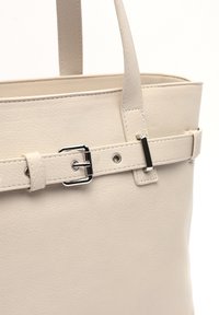 Vaaleanbeige nahkainen käsilaukku, jossa on sileä pinta, jämäkkä muoto ja koristeellinen solkiyksityiskohta sivuhihnassa.