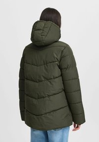 Olivno zelena puhovka s kapuco, z značilnim letevnim šivom in dolgimi rokavi, v kombinaciji s svetlo modrimi jeans hlačami.