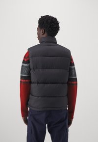 Zwarte gewatteerde vest met horizontale quilted secties, staande kraag, gedragen over een rood patroontrui, gecombineerd met marineblauwe broek.