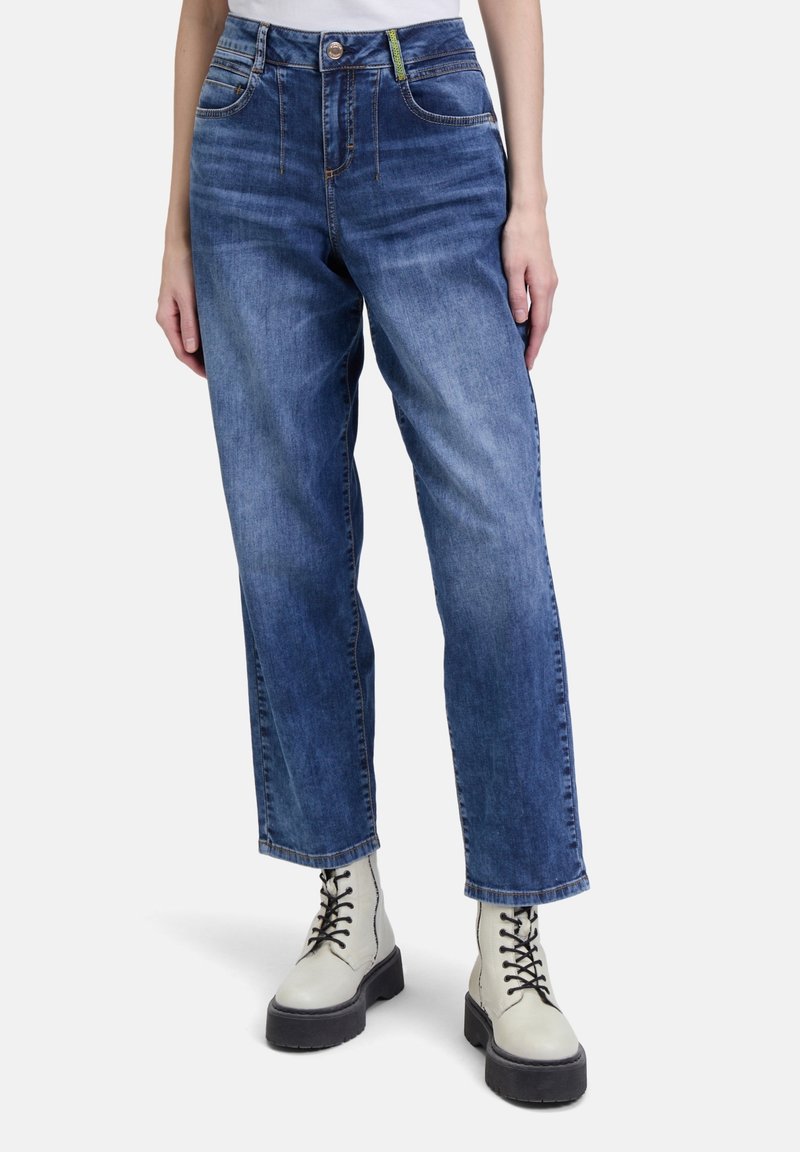 Personne portant un jean bleu droit et des bottes blanches à lacets sur un fond uni, les mains reposant le long du corps.