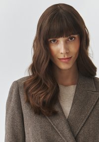 Brauner Blazer mit Fischgrätmuster und Reverskragen, getragen über einem hellbeigen Pullover. Glattes, welliges braunes Haar, das bis zu den Schultern reicht.