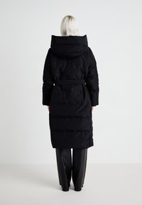 Lange zwarte pufferjas met een capuchon, een ceintuur in de taille en een gestikte uitstraling. Gladde stof en knielange pasvorm, gecombineerd met zwarte broeken.