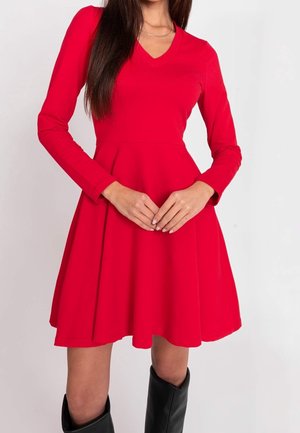 Vrouw draagt een rode jurk met lange mouwen en V-hals, met een uitlopende rok, handen voor zich geklemd, en zwarte kniehoge laarzen.