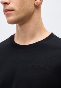 Κοντινό πλάνο του λαιμού και του κάτω μέρους του προσώπου με ασημένιο κρίκο στο αυτί, φορώντας μαύρο πουκάμισο PeakPerformance με κεντημένο λογότυπο στο στήθος.