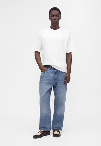 Homme debout, une main dans la poche, portant un t-shirt blanc uni, un jean bleu ample et des mocassins noirs et beiges sur fond blanc.