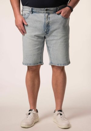 Mand iført lyseblå denimbukser og hvide sneakers mod ensfarvet baggrund, med den ene hånd i lommen og den anden hvilende ved siden.