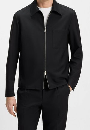 Homme portant une veste noire à manches longues avec fermeture éclair et poche poitrine, sur une chemise blanche et un pantalon noir, main dans la poche.