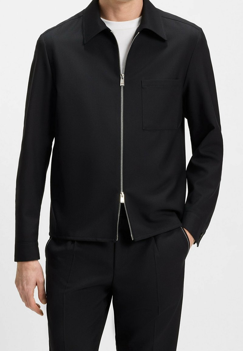 Homme portant une veste noire à manches longues avec fermeture éclair et poche poitrine, sur une chemise blanche et un pantalon noir, main dans la poche.