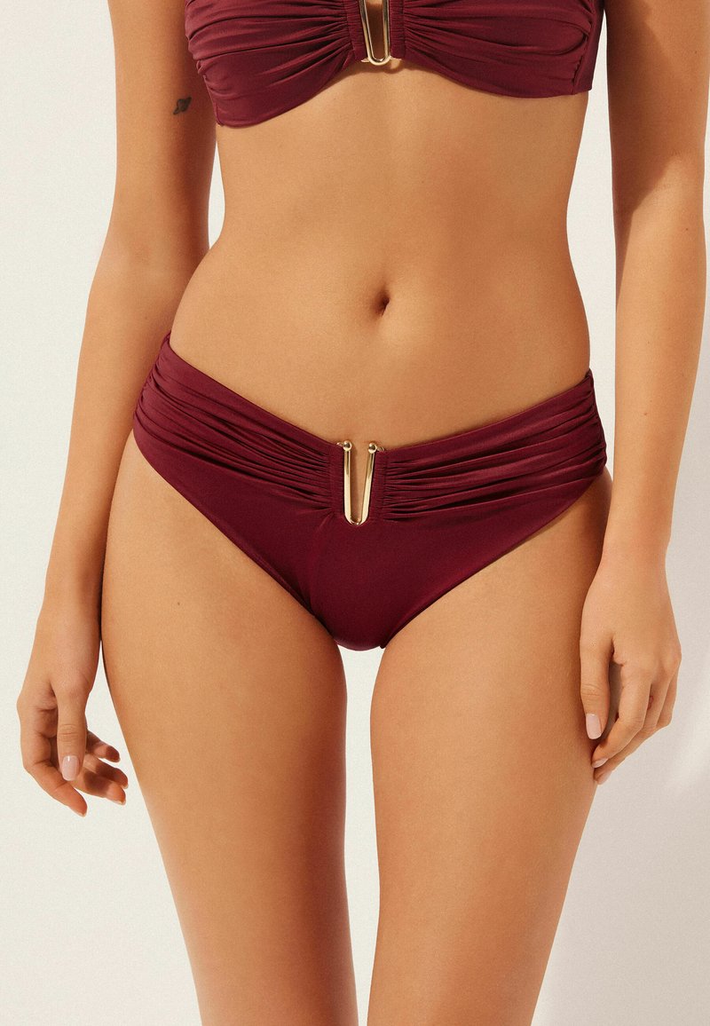 Conjunto de bikini malva que incluye un top fruncido y bragas de talle alto. Detalles en hardware dorado en la parte delantera, textura suave y diseño ceñido al cuerpo.