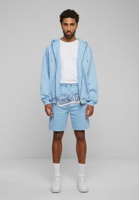 Light blue sweatshirt with zip and pockets, paired with matching shorts featuring white logo. White sneakers and socks complete the outfit.