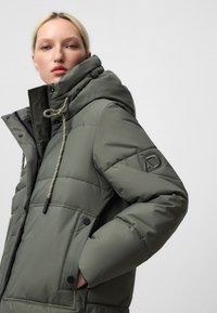 Giacca parka imbottita in verde oliva, con colletto alto, grandi tasche, cappuccio regolabile con cordino e dettaglio logo sulla manica.