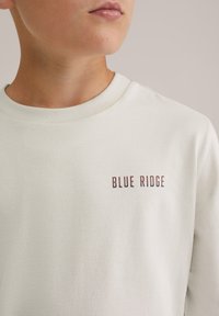 Egy ember világos krémszínű pulóvert visel, amelyen a "BLUE RIDGE" felirat kis, nagybetűs formátumban található a mellrészen.