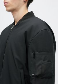 Melnā bomber jaka ar apaļu apkakli, sānu rāvējslēdzēja kabatām un rāvējslēdzēja kabatu uz rokas ar matētu apdari. Gluda auduma tekstūra.