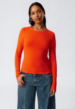 Long-sleeve, fitted top made from a stretchy, smooth fabric in bright orange, paired with loose, blue denim jeans and silver hoop earrings.