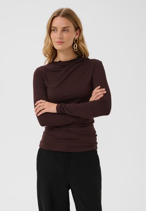 Femme aux cheveux blonds vêtue d'un haut à manches longues marron foncé, d’un pantalon noir, de boucles d'oreilles créoles dorées et de bagues, debout, les bras croisés.