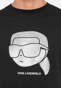 Čierne tričko s trblietavým strieborným grafickým znázornením postavy s okuliarmi a copom, spolu s textom "KARL LAGERFELD" pod ním.