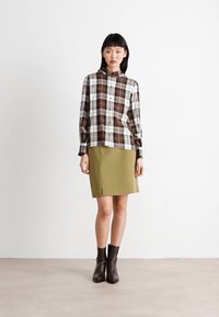 Bruine en witte geruite blouse met een hoge kraag, gecombineerd met een groene rok. Model draagt bruine enkellaarsjes. Vaak stof met een gestructureerde pasvorm.