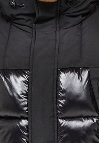 Svart puffjacka med matt finish och glänsande paneldetaljer. Har dragkedja, huva och horisontella bafflar för isolering.